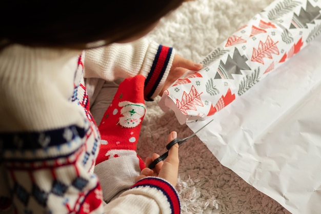 Mujer joven en casa empacando regalos para Navidad y año nuevo, cortando papel con tijeras