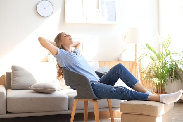 Mujer joven en casa sentada en una silla moderna frente a la ventana, relajándose en su sala de estar.