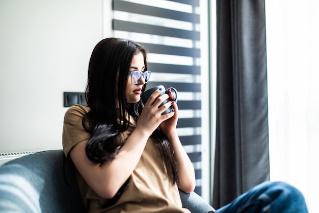 Mujer joven en casa sentado en una silla moderna delante de la ventana relajándose en su sala de estar bebiendo café