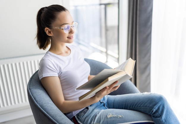 Mujer joven en casa sentado en una silla moderna delante de la ventana relajándose en su sala de estar leyendo el libro