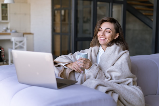 Mujer joven en casa en un sofá en invierno bajo una acogedora manta con un ordenador portátil, viendo series de comedia