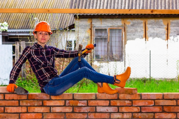 Una mujer joven en un casco de construcción con un martillo de construcción y una paleta en sus manos está sentada en una pared de ladrillos