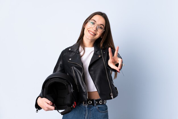 Mujer joven con un casco de moto sobre pared azul aislado sonriendo y mostrando el signo de la victoria