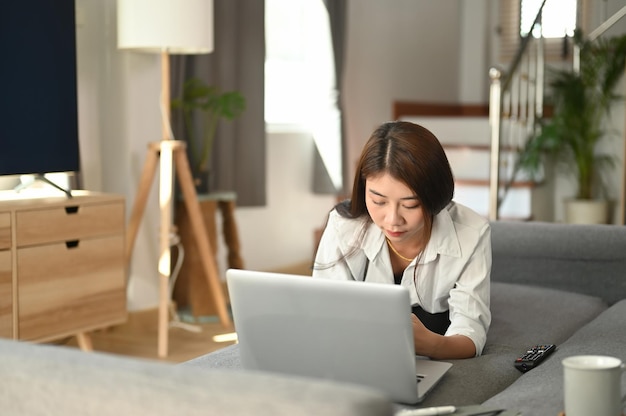 Mujer joven casual acostada en el sofá y usando una computadora portátil