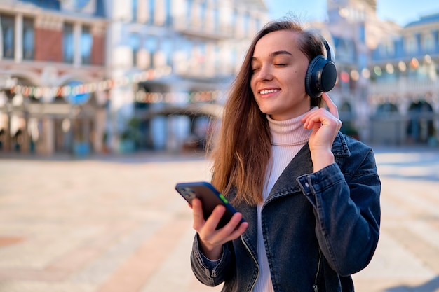 Mujer joven casual feliz sonriente moderna que usa auriculares inalámbricos y smartphone para escuchar música al aire libre