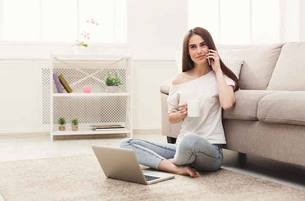 Mujer joven en casual sentada en el suelo hablando por el móvil usando la computadora portátil, bebiendo té. Copia espacio