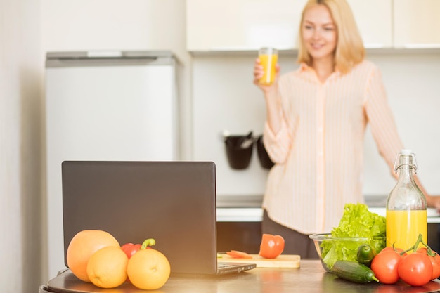 Mujer joven caucásica con camisa a rayas sosteniendo un vaso de jugo fresco de pie en la cocina detrás de la mesa con computadora portátil, verduras y frutas, viendo el espectáculo mientras se cocina.