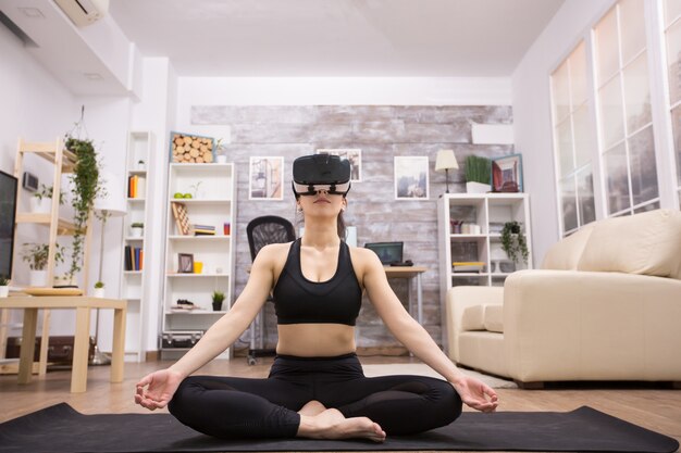 Mujer joven caucásica haciendo yoga en la sala de estar y con gafas de realidad virtual.