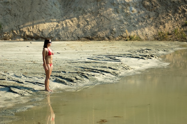 Mujer joven caucásica hermosa delgada en bikini y gafas de sol se encuentra en la orilla arenosa cerca del lago