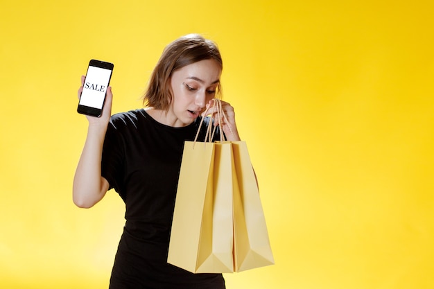 Mujer joven caucásica sonriendo y sosteniendo el teléfono móvil y el paquete de compras y mirando a la pantalla del teléfono