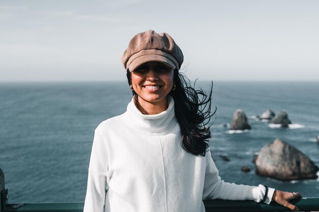 Mujer joven caucásica con suéter blanco y sombrero marrón sonriendo mirando a la cámara apoyándose en el