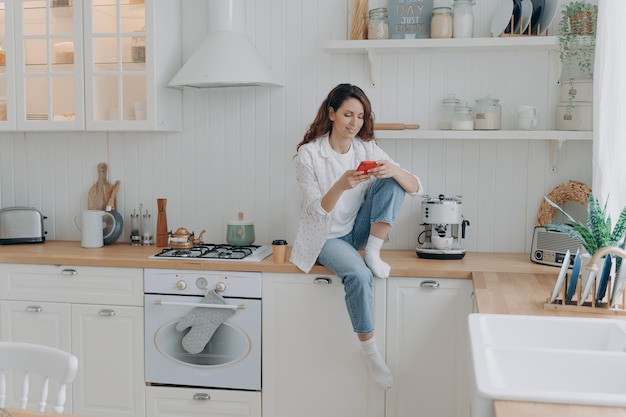 Mujer joven caucásica con teléfono sentado en la encimera de la cocina y enviando mensajes de texto Concepto de estilo de vida