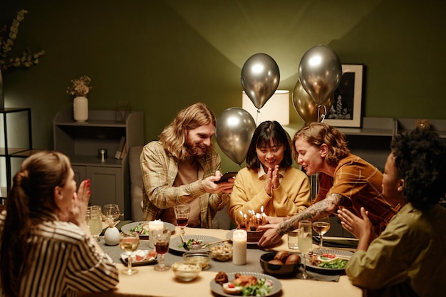 Mujer joven celebrando un cumpleaños con sus amigos sentados en la mesa del comedor con pastel de cumpleaños en la sala de estar