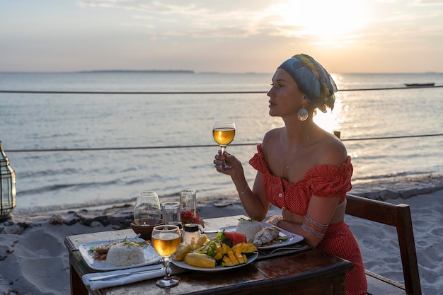 Mujer joven con una cena romántica en el restaurante del hotel durante la puesta de sol cerca de las olas del mar en la playa tropical de cerca