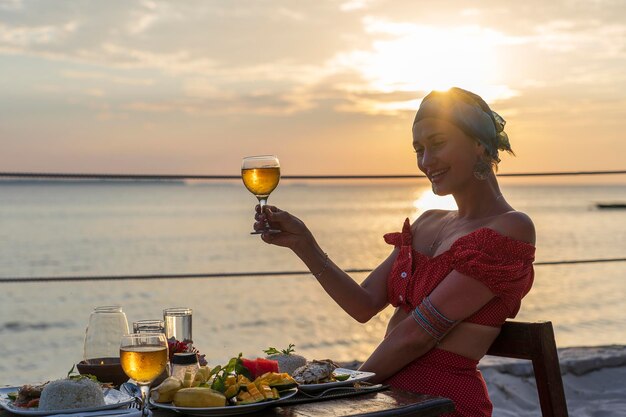 Mujer joven con una cena romántica en el restaurante del hotel durante la puesta de sol cerca de las olas del mar en la playa tropical de cerca