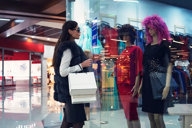 Mujer joven en el centro comercial de pie cerca del escaparate y mirando dos maniquíes con vestidos y pelucas. Chica atractiva de moda está haciendo compras en el centro comercial. Señora con gafas en busca de atuendo.