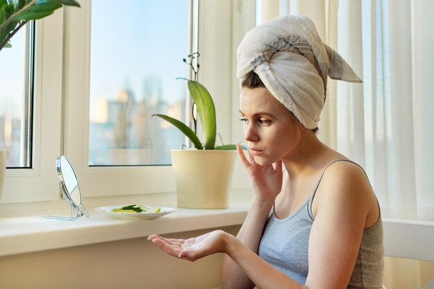 Mujer joven cerca de la ventana de casa mirándose en el espejo aplicando crema facial