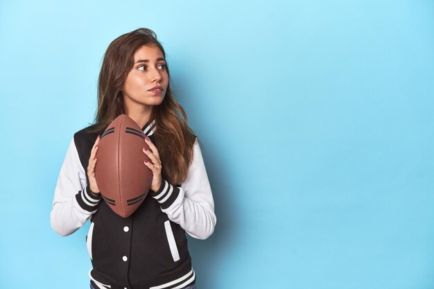 Foto mujer joven con chaqueta de béisbol con pelota de fútbol en el estudio