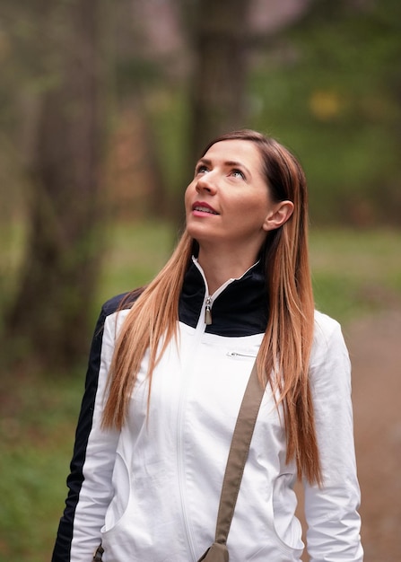 Mujer joven con chaqueta deportiva blanca camina en el bosque, mira hacia el cielo, árboles borrosos detrás de ella