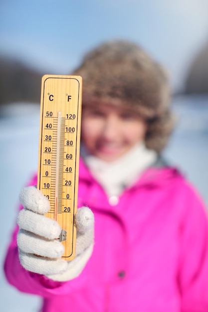 Foto mujer joven con chaqueta deportiva rosa sosteniendo un termómetro de madera afuera en un día frío, ilustrando el clima con una temperatura tan baja como -20 grados centígrados.