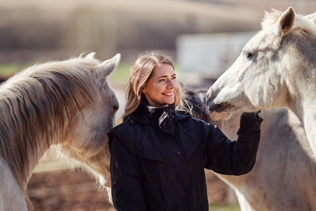 Mujer joven con chaqueta de equitación negra de pie cerca de un grupo de caballos árabes blancos sonriendo feliz uno en cada lado detalle de primer plano