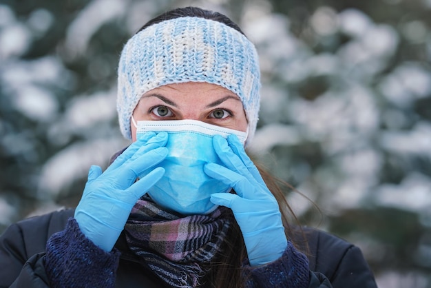 Mujer joven en chaqueta de invierno con máscara de virus facial desechable azul de un solo uso y guantes protectores, fondo borroso de árboles cubiertos de nieve, detalle de cierre