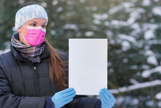 Mujer joven con chaqueta de invierno con máscara de virus facial rosa desechable de un solo uso y guantes protectores, sosteniendo una mesa blanca vacía - espacio para texto - mirándola, fondo borroso de árboles cubiertos de nieve