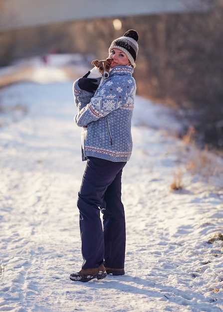 Mujer joven en chaqueta de invierno que lleva su Jack Russell terrier en las manos - enfoque en la cara del perro, fondo borroso de carretera de campo cubierto de nieve