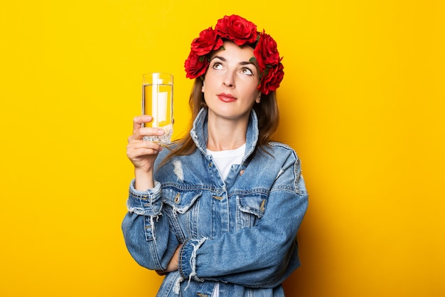 Mujer joven en chaqueta de mezclilla y una corona de flores rojas en la cabeza sosteniendo un vaso de agua sobre una pared amarilla.