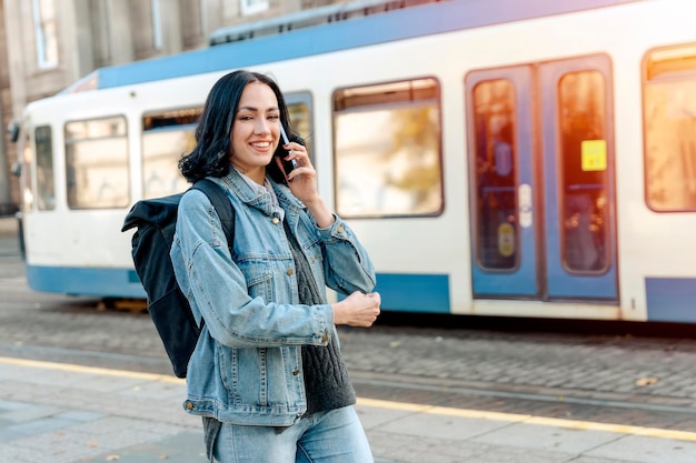 Una mujer joven con una chaqueta de mezclilla está hablando por teléfono y esperando un tranvía en la parada