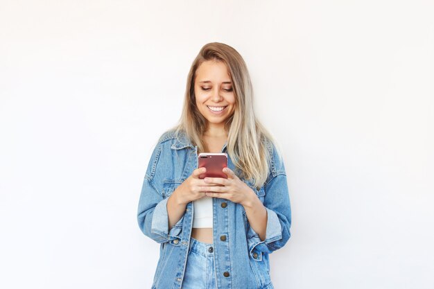 Una mujer joven con una chaqueta de mezclilla y jeans sonríe y sostiene un teléfono móvil mirando la pantalla