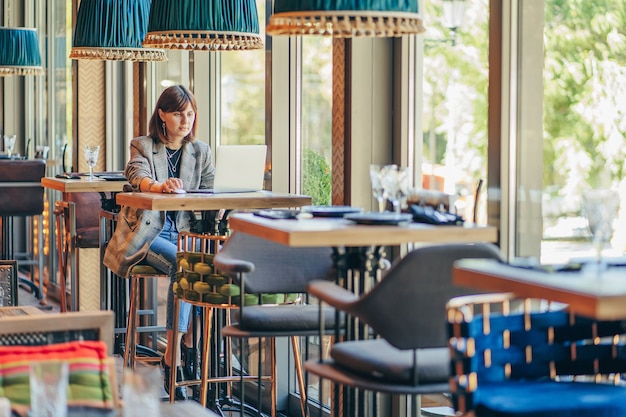 Mujer joven en chaqueta con portátil en el café cerca de la ventana. professions es bloguero, autónomo y escritor. freelancer que trabaja en la cafetería. aprendizaje en línea.