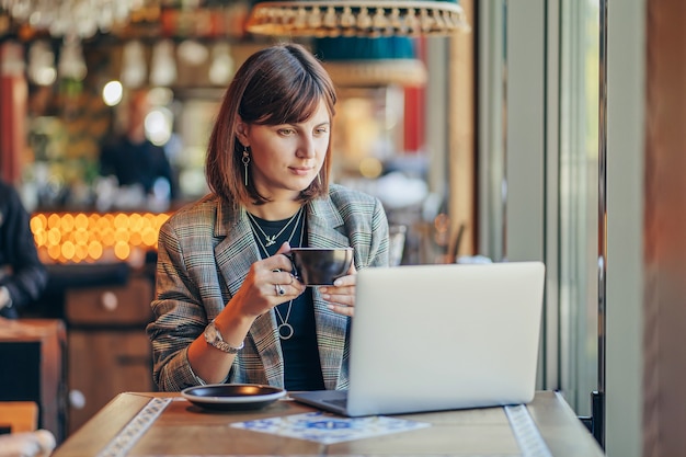 Mujer joven en chaqueta con portátil en el café cerca de la ventana. Professions es bloguero, autónomo y escritor. Freelancer que trabaja en la cafetería. Aprendizaje en línea.