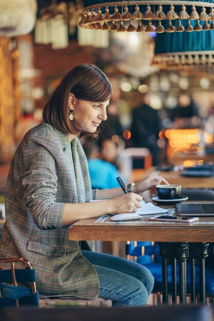 Mujer joven en chaqueta con portátil en el café cerca de la ventana. Professions es bloguero, autónomo y escritor. Freelancer que trabaja en la cafetería. Aprendizaje en línea.