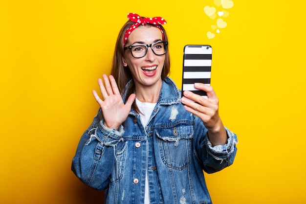 Mujer joven con chaqueta vaquera y gafas sostiene un teléfono en sus manos y habla por video chat en una pared amarilla.