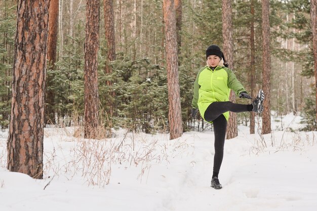 Mujer joven en chaqueta verde de pie en el bosque de invierno y levantando la pierna a un lado en la formación