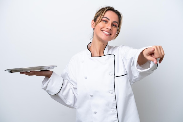 Mujer joven chef con bandeja aislada sobre fondo blanco apuntando al frente con expresión feliz