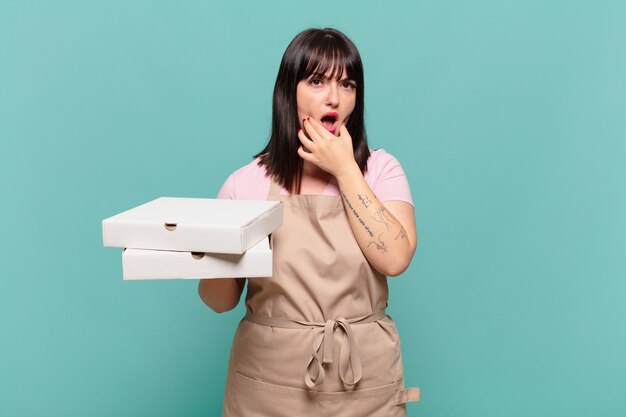 Mujer joven chef con la boca y los ojos bien abiertos y la mano en la barbilla, sintiéndose desagradablemente conmocionada, diciendo qué o guau