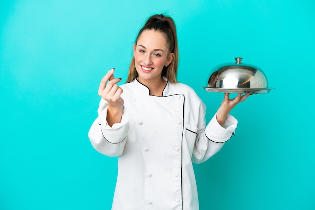 Mujer joven chef caucásica con bandeja aislada sobre fondo azul haciendo gesto de dinero