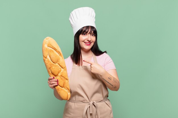 Mujer joven chef sonriendo alegremente, sintiéndose feliz y apuntando hacia un lado y hacia arriba, mostrando el objeto en el espacio de la copia