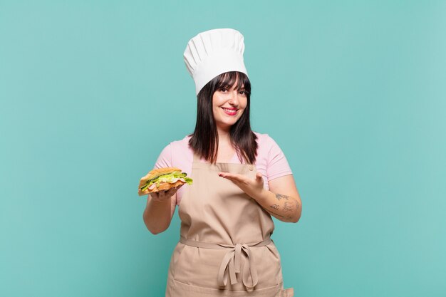 Mujer joven chef sonriendo alegremente, sintiéndose feliz y mostrando un concepto en el espacio de la copia con la palma de la mano