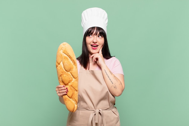 Mujer joven chef sonriendo felizmente y soñando despierto o dudando, mirando hacia el lado