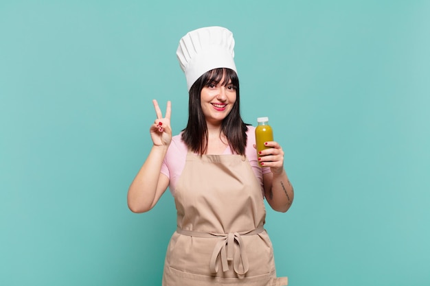 Mujer joven chef sonriendo y mirando amistosamente, mostrando el número dos o el segundo con la mano hacia adelante, contando hacia atrás