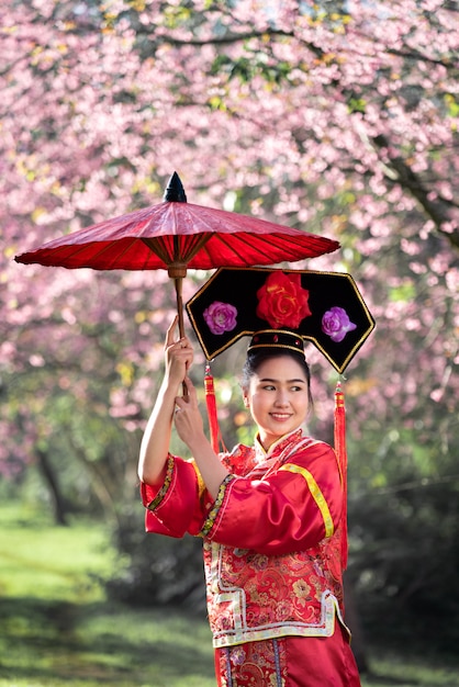 Mujer joven china hermosa que lleva el cheongsam tradicional rojo en jardín de los cerezos en flor