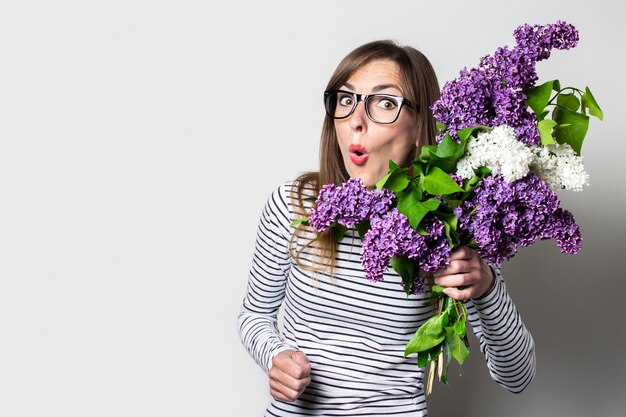 La mujer joven del choque en vidrios sostiene un ramo de lilas en un fondo claro.