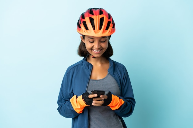 Mujer joven ciclista aislada en la pared azul enviando un mensaje con el móvil