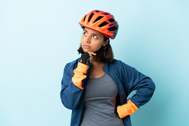 Mujer joven ciclista aislada en la pared azul que tiene dudas y con expresión de la cara confusa