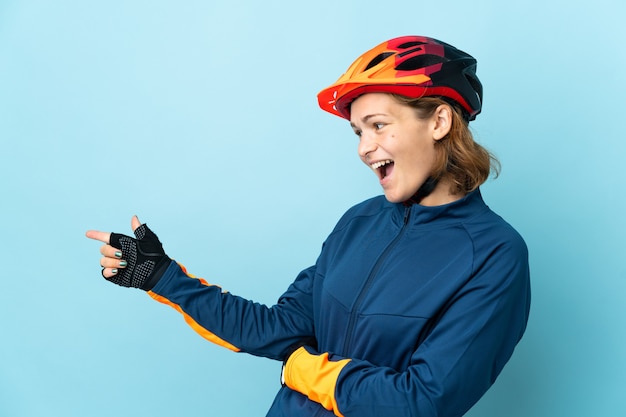 Mujer joven ciclista aislada sobre fondo azul apuntando con el dedo hacia el lado y presentando un producto