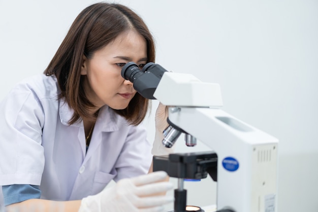 Una mujer joven científica con una bata de laboratorio mirando a través de un microscopio en un laboratorio para investigar y experimentar. Científico que trabaja en un laboratorio. Educación foto de stock