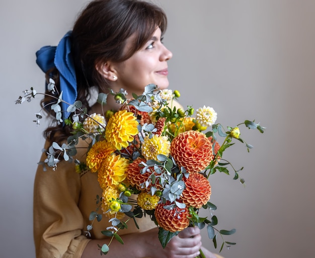 Mujer joven con una cinta azul en el pelo, sosteniendo un ramo de crisantemos amarillos y naranjas, fondo gris.